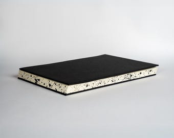 May include: A rectangular notebook with a solid black cover and cream-colored pages. The edges of the paper block feature a distinct black speckled pattern, creating a high-contrast visual effect against the plain black front cover.