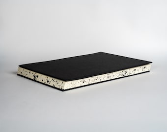 May include: A rectangular notebook with a solid black cover and cream-colored pages. The edges of the paper block feature a distinct black speckled pattern, creating a high-contrast visual effect against the plain black front cover.