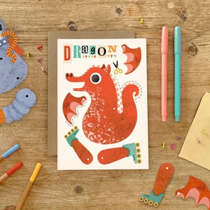 May include: A paper craft kit featuring a red dragon with wings, legs, and a tail. The dragon is on a white card with the word "DRAGON" printed in colorful letters. The kit includes a pair of scissors and a dotted line to cut out the pieces.