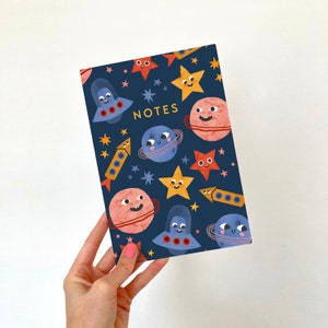 Op de afbeelding: Een blauw notitieboek met een ruimtethema. De omslag heeft planeten, sterren, raketten en ruimteschepen met lachende gezichten. De tekst "NOTES" is in geel op de omslag gedrukt.