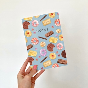 Puede incluir: Una libreta azul con la palabra "NOTES" impresa en la portada. La portada presenta un patrón repetido de varias galletas y bizcochos, incluyendo chocolate, jengibre y galletas de mantequilla.