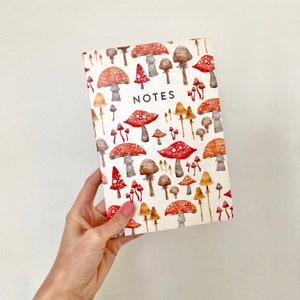 Op de afbeelding: Een wit notitieboek met een paddenstoelmotief. De omslag heeft rode en bruine paddenstoelen met witte stelen. De tekst "NOTES" is in zwart op de omslag gedrukt.