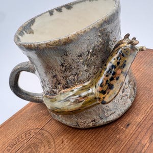 May include: A handcrafted ceramic mug with a distinctive design. The mug features a mottled grey exterior, a cream interior, and a sculpted slug attached to the side. The slug is brown, tan, and black. The mug has a handle and rests on a wooden surface.