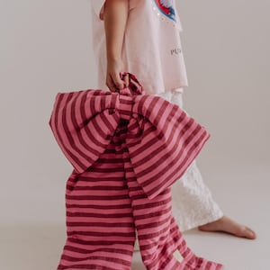 May include: A large, oversized fabric bow in alternating horizontal stripes of light pink and dark maroon. The bow features a central knot and two long, rectangular fabric tails hanging downward, with a small rectangular label on one tail.