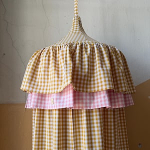 May include: A fabric canopy featuring a mustard yellow and white gingham pattern. The top section has a conical shape with a gathered, double-layered ruffle trim. The upper ruffle is mustard yellow gingham, and the lower ruffle is light pink gingham.