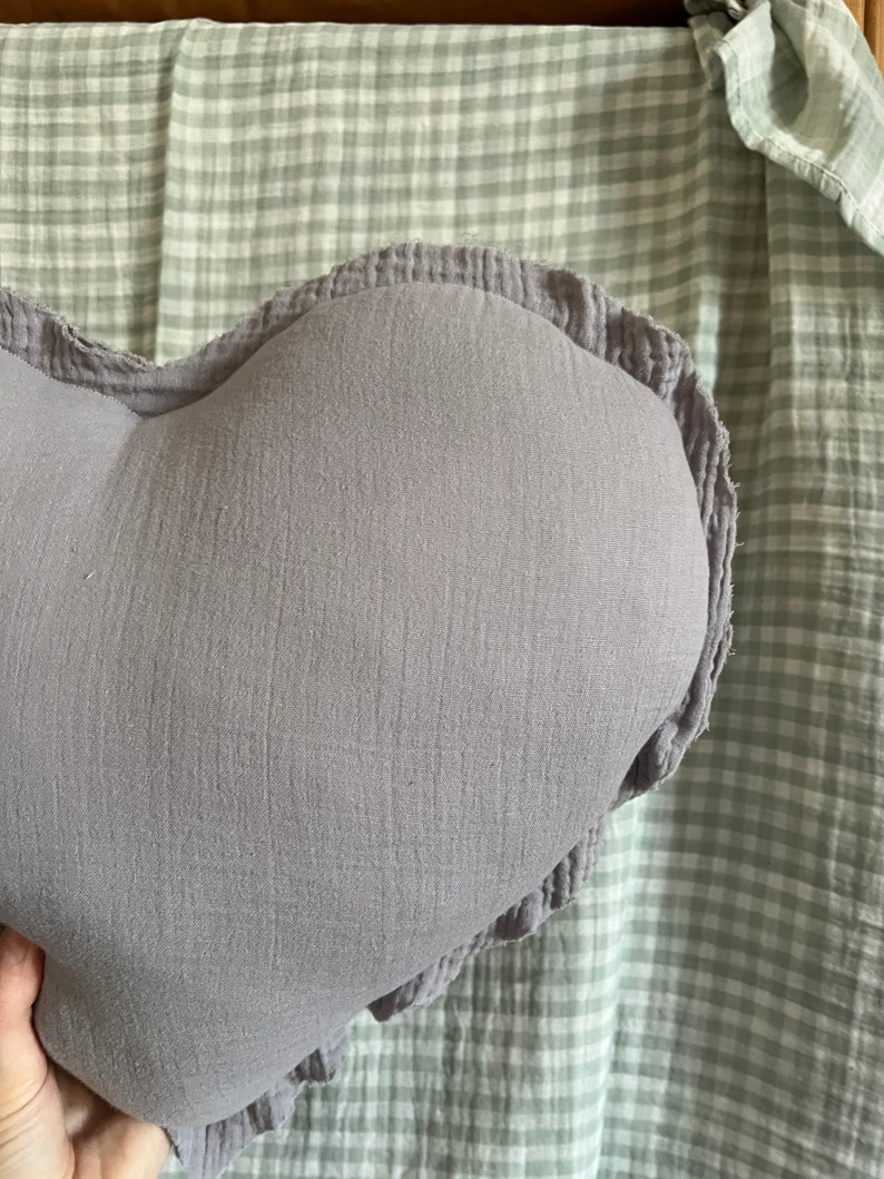 May include: A soft, heart-shaped pillow with a ruffled edge. The pillow is made of a light purple fabric.