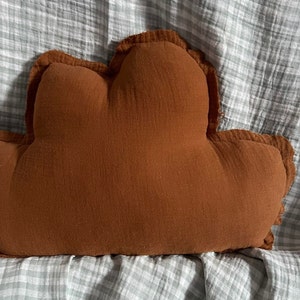 May include: A brown cloud-shaped pillow with a soft, textured fabric. The pillow is on a gray and white checkered surface.