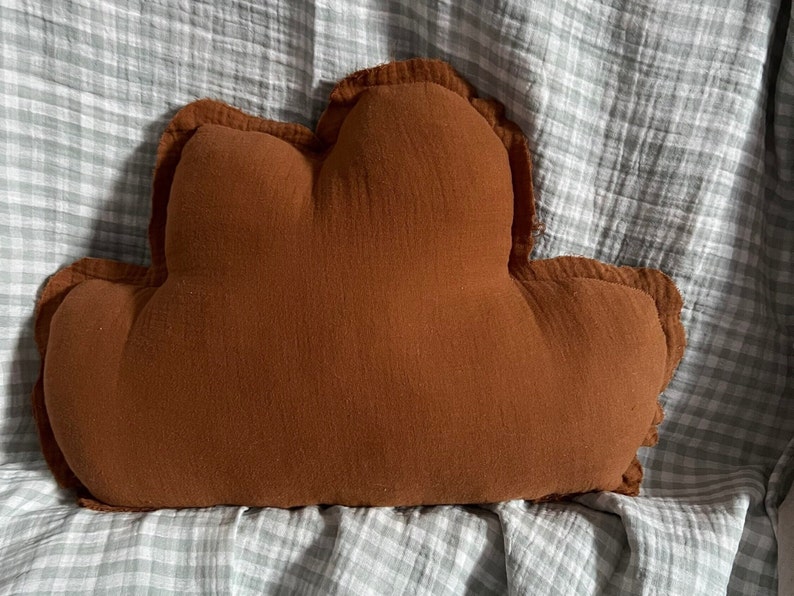 May include: A brown cloud-shaped pillow with a soft, textured fabric. The pillow is on a gray and white checkered surface.