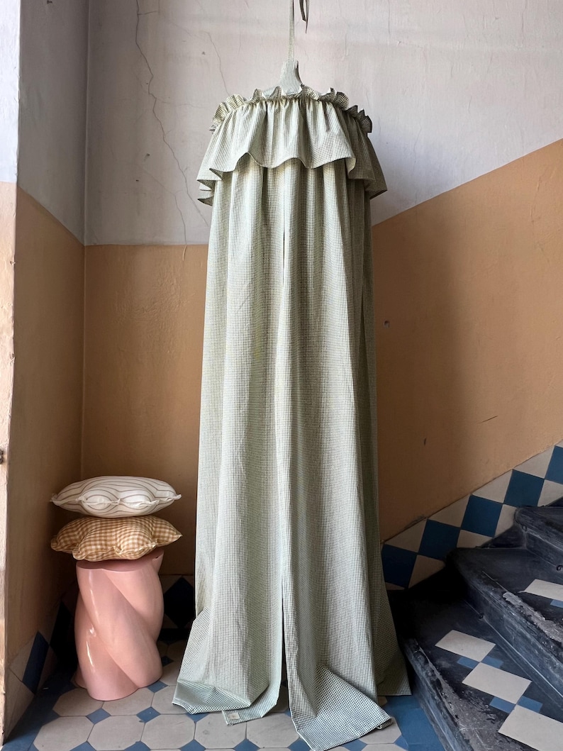 May include: A green and white gingham canopy with a ruffled top hangs from the ceiling in a room with a pink pedestal and patterned pillows.