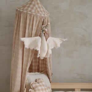 May include: A beige and white gingham canopy hangs over a bed with a white duvet. A plush white goose with outstretched wings is suspended from the canopy. A matching gingham pillow and a wooden toy car are on the bed.