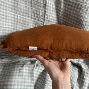 May include: A brown, rectangular pillow with a white label that says "ALO" on it. The pillow is made of a soft, textured fabric and has a slightly curved shape.