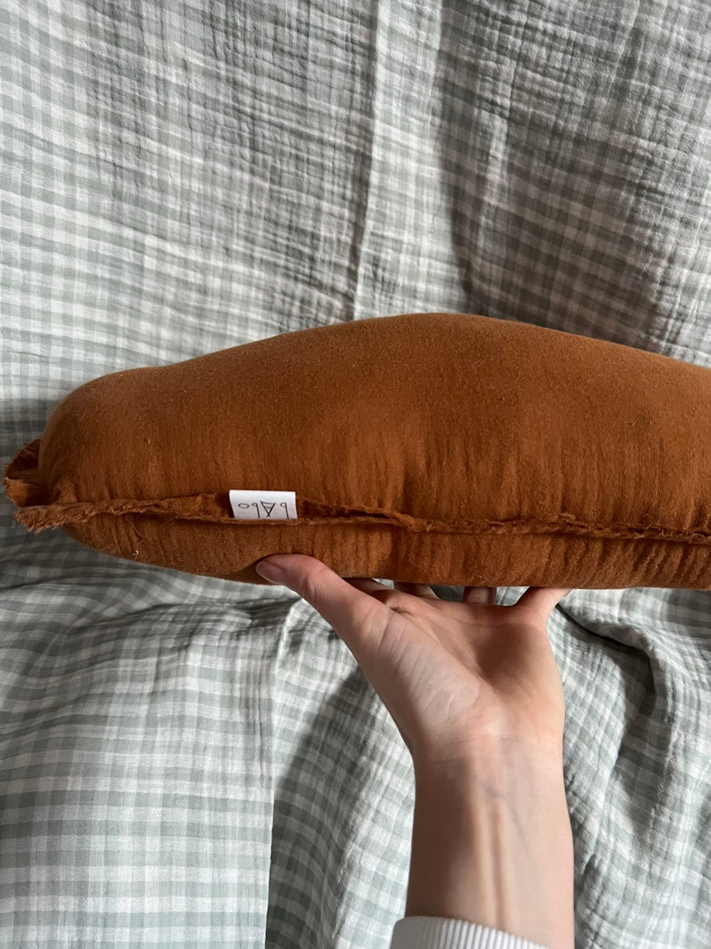 May include: A brown, rectangular pillow with a white label that says "ALO" on it. The pillow is made of a soft, textured fabric and has a slightly curved shape.