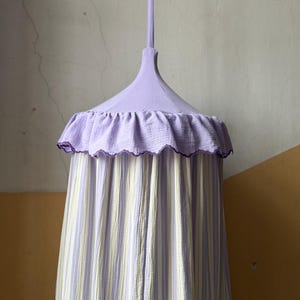 May include: A lavender-colored canopy with a ruffled edge and striped fabric. The canopy has a scalloped trim and a hanging loop at the top. The fabric has vertical stripes of lavender, white, and pale yellow.