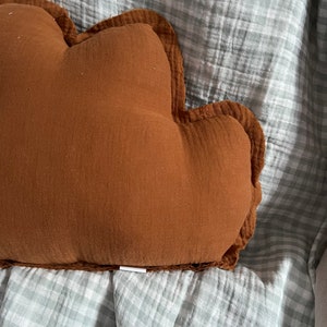 May include: A brown cloud-shaped pillow with a ruffled edge, resting on a light blue and white gingham fabric.