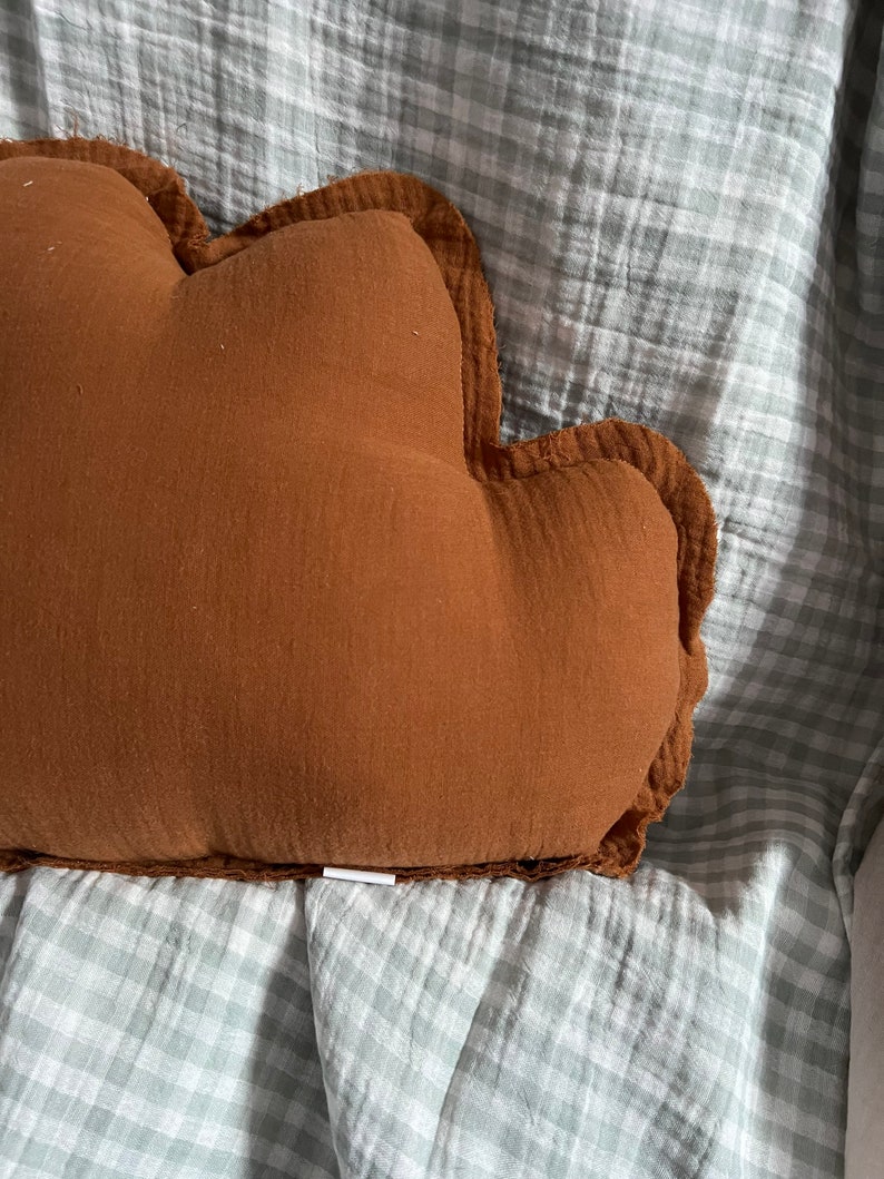 May include: A brown cloud-shaped pillow with a ruffled edge, resting on a light blue and white gingham fabric.