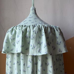 May include: A pale green canopy featuring a delicate pattern of tiny purple flowers. The canopy has a ruffled trim and a conical top, suitable for a bed or play space. The fabric looks to be a soft, textured material.