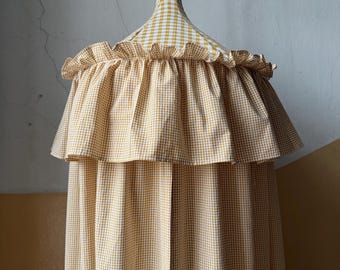 May include: A yellow and white gingham patterned canopy with a ruffled edge. The canopy is suspended from a matching gingham fabric-covered pole. The fabric is a light, airy material, suitable for a nursery or children's room.