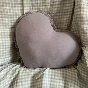 May include: A heart-shaped pillow with a soft, gray fabric and ruffled edges. The pillow is sitting on a white and gray checkered fabric.