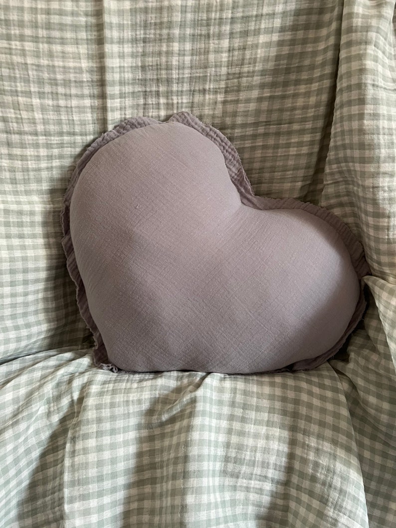 May include: A heart-shaped pillow with a soft, gray fabric and ruffled edges. The pillow is sitting on a white and gray checkered fabric.