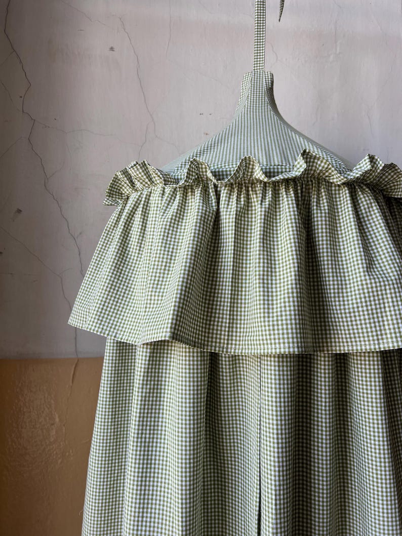 May include: A green and white gingham canopy with a ruffled edge. The canopy is suspended from a white ceiling.