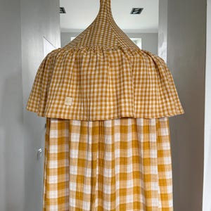 Può includere: Un baldacchino a quadretti giallo senape e bianco, caratterizzato da una parte superiore conica con un occhiello per appenderlo e una gonna a balze. Il tessuto ha un aspetto testurizzato e stropicciato, con una piccola etichetta beige.