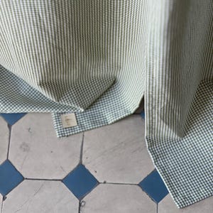 May include: Green and white gingham fabric with a small tag attached. The fabric is folded and draped over a tiled floor.