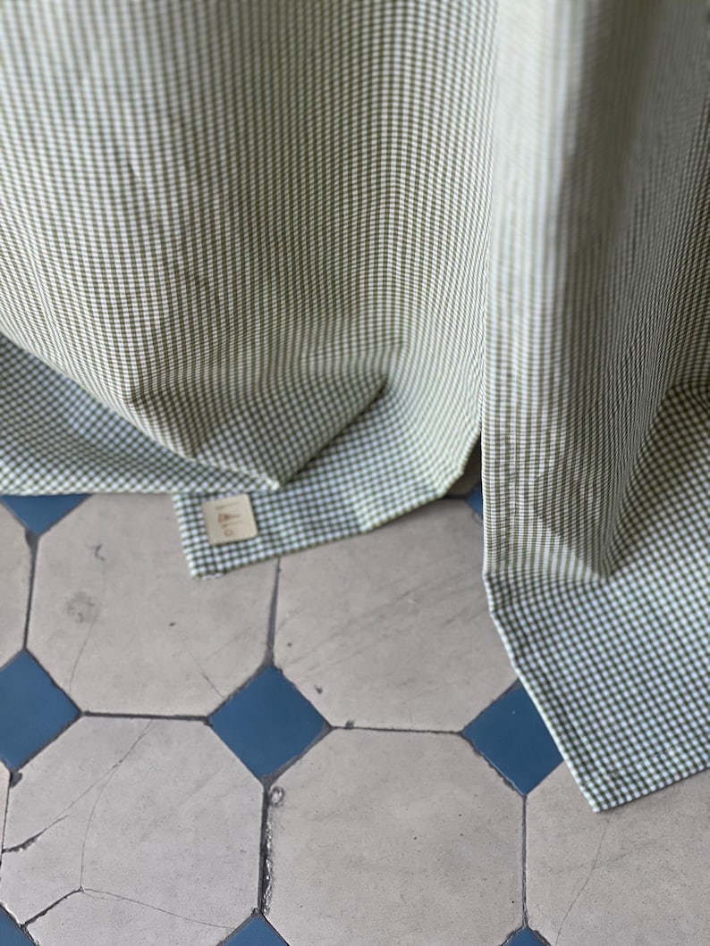 May include: Green and white gingham fabric with a small tag attached. The fabric is folded and draped over a tiled floor.