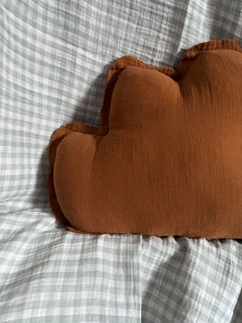 May include: A brown cloud-shaped pillow with a ruffled edge, resting on a white and gray gingham fabric.