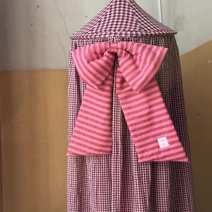 Puede incluir: Un dosel de tela cónico con un patrón de cuadros vichy en granate y blanco. Un lazo decorativo grande hecho de tela a rayas horizontales rosas y granates está sujeto al frente. Se observa una pequeña etiqueta rectangular blanca con texto.