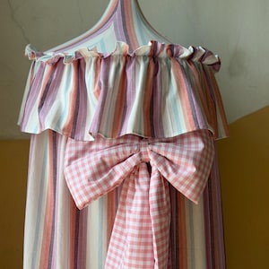May include: A conical fabric canopy featuring vertical stripes in shades of pink, purple, orange, and light blue. The top edge has a ruffled trim, and a large, pink and white gingham check fabric bow is attached to the front with long streamers.