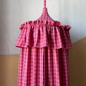 Pode incluir: Um dossel com padrão xadrez vichy em rosa e rosa escuro, apresentando uma secção superior cónica com uma alça para pendurar e um folho largo e franzido em torno da circunferência superior. O tecido exibe um padrão xadrez consistente.
