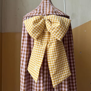 May include: A conical fabric canopy featuring a brown and white gingham pattern. Attached to the front is a large, decorative bow made from a yellow and white gingham fabric, which hangs down with two long, pointed tails over the canopy.