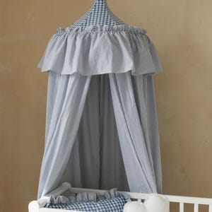 May include: A blue and white striped canopy hangs over a white crib. The canopy has a ruffled edge and a gingham top. A gingham pillow and a cloud-shaped pillow are in the crib.