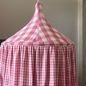 May include: A pink and white gingham canopy for a bed or play area. The canopy is made of a soft, lightweight fabric and has a ruffled edge.