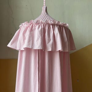 May include: A pink and white striped canopy with a ruffled edge. The canopy is suspended from a matching striped fabric tie. The top of the canopy is a pink and white gingham pattern.