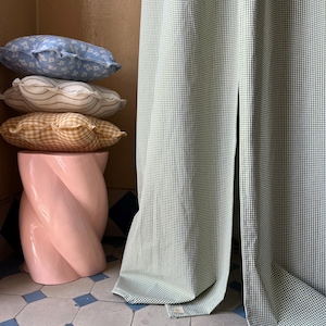 May include: A stack of three decorative pillows in various patterns and colors, including blue, white, and yellow, are placed on a pink, twisted pedestal. A green and white gingham curtain is draped behind the pillows.