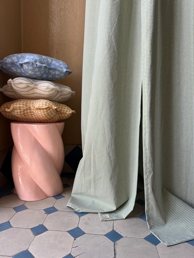 May include: A stack of three decorative pillows in various patterns and colors, including blue, white, and yellow, are placed on a pink, twisted pedestal. A green and white gingham curtain is draped behind the pillows.