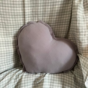 May include: A heart-shaped pillow with a ruffled edge, made of a soft, light gray fabric. The pillow is sitting on a white and gray checkered blanket.