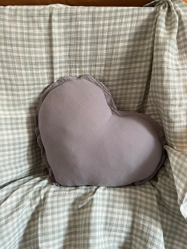 May include: A heart-shaped pillow with a ruffled edge, made of a soft, light gray fabric. The pillow is sitting on a white and gray checkered blanket.