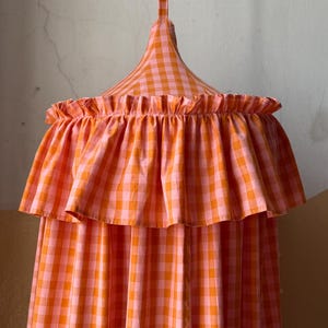Op de afbeelding: Een hangende luifel in een levendig oranje en roze ruitpatroon. De luifel heeft een gefranjerde rand en een conische top, wat een speels en decoratief element creëert. De stof is lichtgewicht en lijkt katoen of een vergelijkbaar materiaal te zijn.