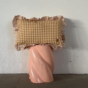 May include: A yellow and white gingham pillow with a ruffled edge sits on a pink twisted pedestal.