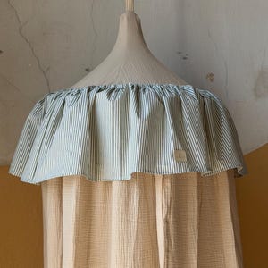 May include: A beige fabric canopy featuring a gathered top ruffle with a green and white vertical stripe pattern. The main body of the canopy is made of a textured, solid beige material. A small rectangular fabric label is attached to the ruffle.