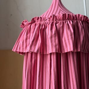 Könnte beinhalten: Ein rosa-braun gestreifter Baldachin mit gerüschtem Rand. Der Baldachin hat eine konische Spitze und eine Aufhängeschlaufe. Der Stoff ist weiche Baumwolle und die Streifen sind vertikal. Das Gesamtdesign ist skurril und dekorativ.