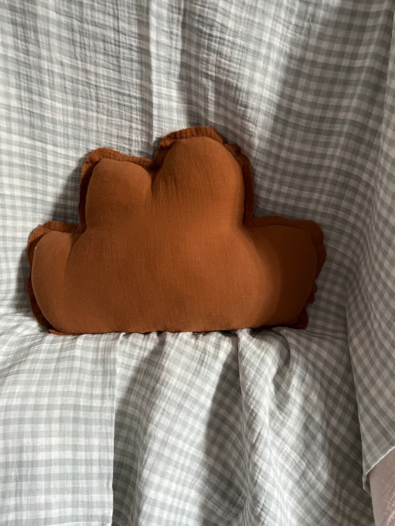 May include: A brown cloud-shaped pillow with a ruffled edge sits on a gray and white gingham fabric.