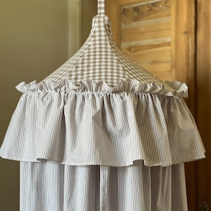 Puede incluir: Un dosel a rayas beige y blanco con un borde con volantes. La parte superior del dosel presenta un patrón de cuadros beige y blanco. El dosel cuelga de un lazo de tela a juego.