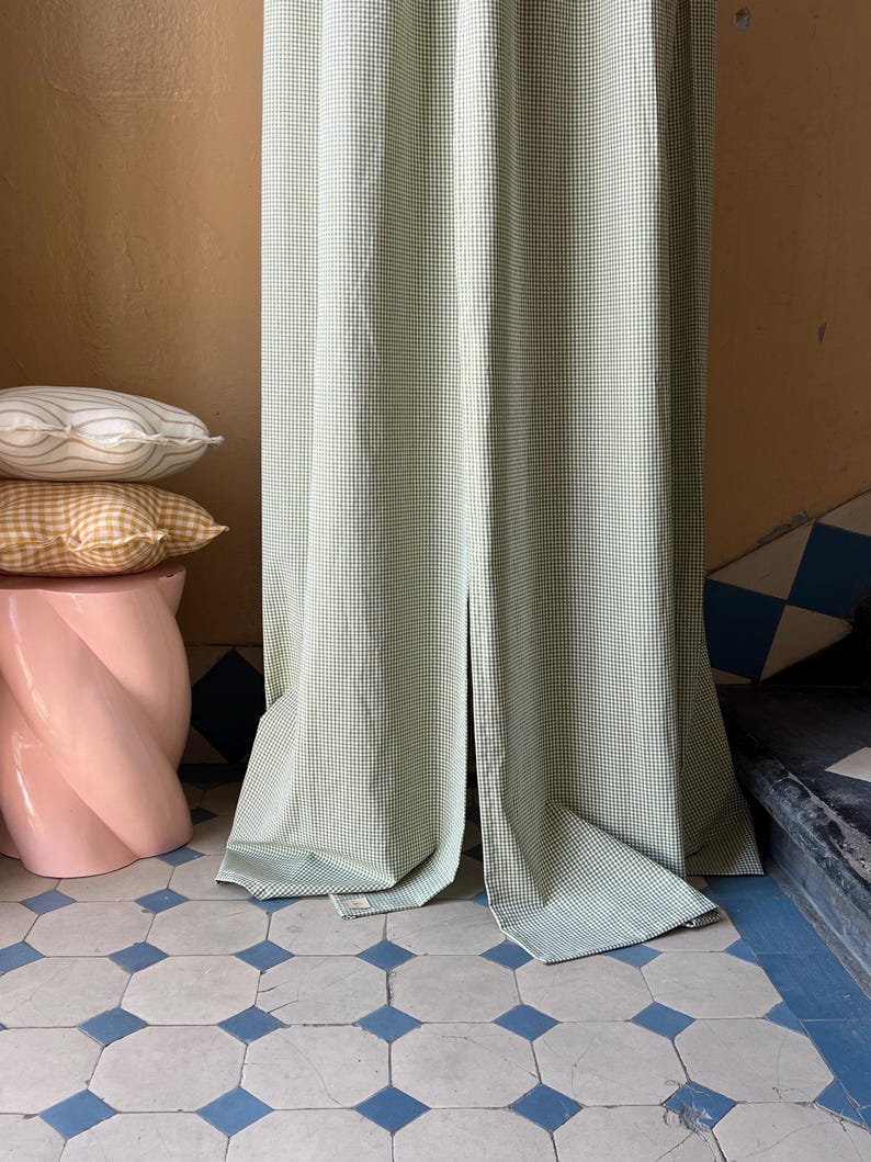 May include: A green and white gingham curtain hangs in a doorway with a pink pedestal and patterned pillows in the foreground. The floor is tiled in a white and blue geometric pattern.