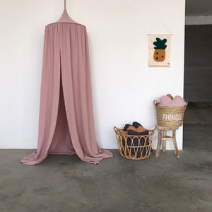 Puede incluir: Una tienda de campaña de dosel rosa polvoriento cuelga del techo en una habitación con un tapiz de pared de piña tejido y una cesta de mimbre llena de almohadas. La cesta se coloca sobre un pequeño taburete de madera con la palabra "THINGS" impresa en él.