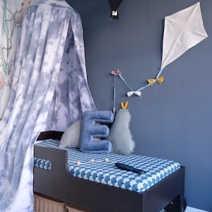 Puede incluir: Un dosel azul y blanco tie-dye sobre un marco de cama de madera oscura con un colchón estampado en azul y blanco. La cama está decorada con una letra E azul y una almohada gris y blanca.