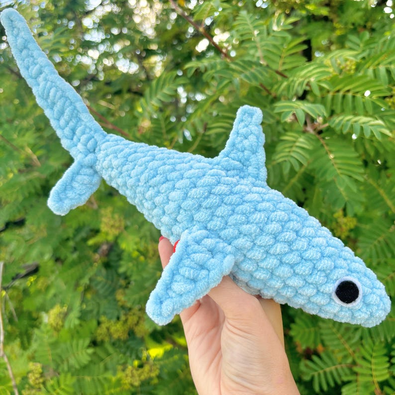 Pu&ograve; includere: Un giocattolo di squalo di peluche di colore azzurro chiaro lavorato a uncinetto con occhi neri. Il giocattolo &egrave; tenuto in una mano su uno sfondo verde di foglie.