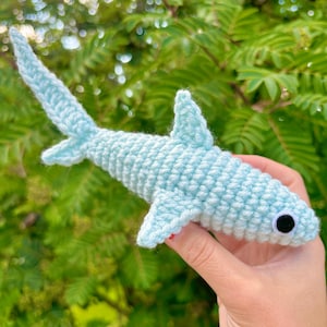 Pu&ograve; includere: Un giocattolo a forma di squalo martello in crochet di colore azzurro chiaro con un occhio nero. Il giocattolo &egrave; tenuto da una mano.